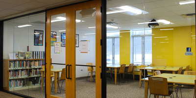 The Hub room with double glass doors and glass windows. There are tables and chairs and a bookcase in the room.